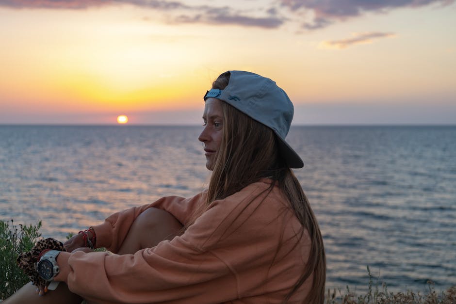 A woman in a cap enjoys a serene sunset by the ocean, showcasing relaxation and natural beauty.