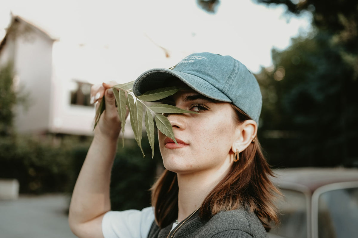 A confident young woman with a baseball cap holds a leaf outdoors, blending nature and fashion.