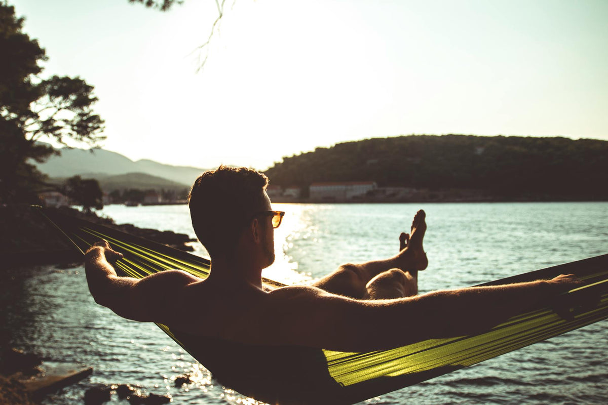 A man relaxes in a hammock by a tranquil lake during sunset, enjoying a peaceful moment.