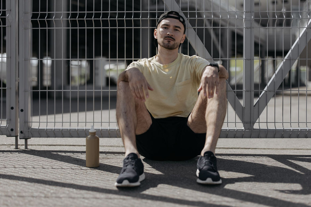 Casually dressed young man sitting by a fence with a bottle, enjoying a sunny day.
